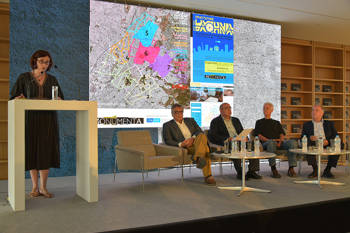 A woman makes a speech on the podium and four men sitting on the stage in the form of a panel watch her